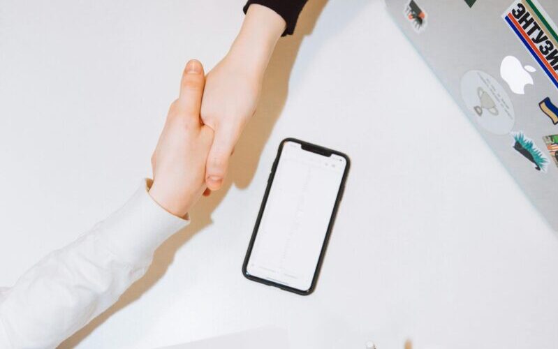 Due persone si stringono la mano con un iPhone e un MacBook sul tavolo.